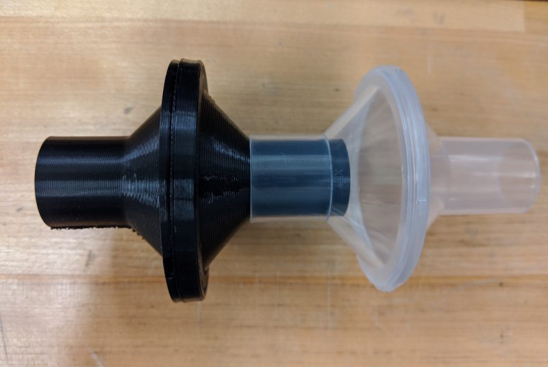 A black 3-D printed part is connected to a clear part. Each part widens in the middle where there's room for a filter to be inserted.