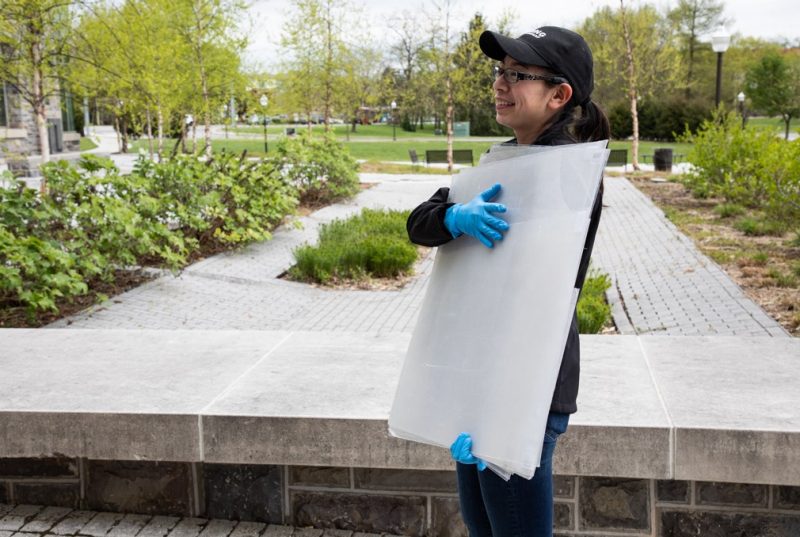 A person wearing a black cap and blue plastic gloves holds sheets of plastic.